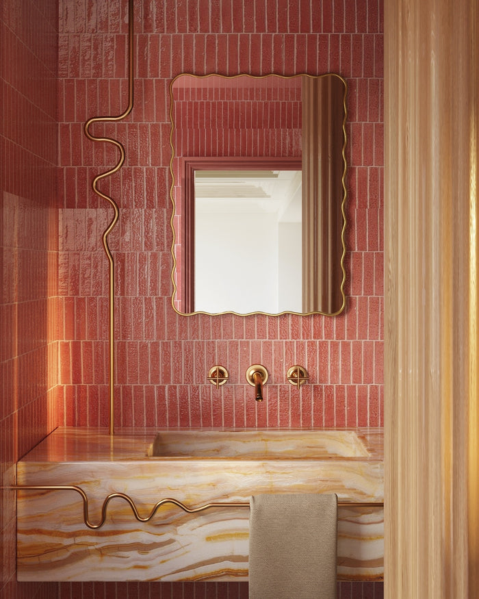 Rectangular wall mirror with a softly wavy gold frame mounted on a pink tiled wall above a marble bathroom sink with brass fixtures.