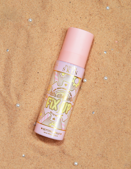 A bottle of P.Louise Fix Up Mattifying Setting Spray with a pink cap and decorative patterns on the label, displayed on a sandy surface.